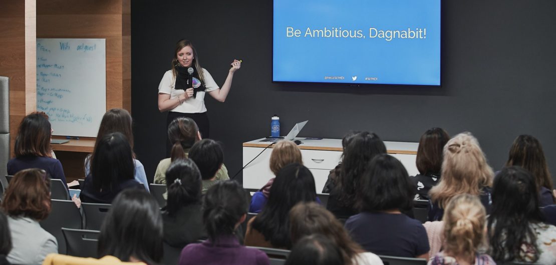 Holly stands in front of screen encouraging audience to "be ambitious!"